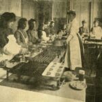 A woman stands before, and teaching, a group of women who work over table-top grills and cook pots.