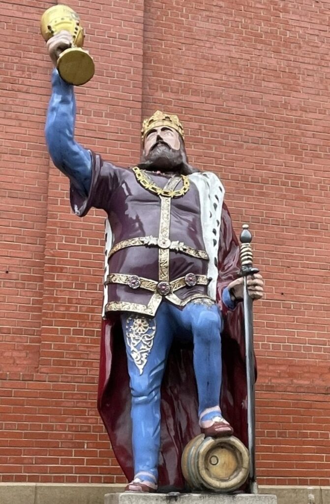 A statue of a king with a sword in one hand, the other hand raises a chalice into the air, he stands with one leg bent, foot resting on a barrel.