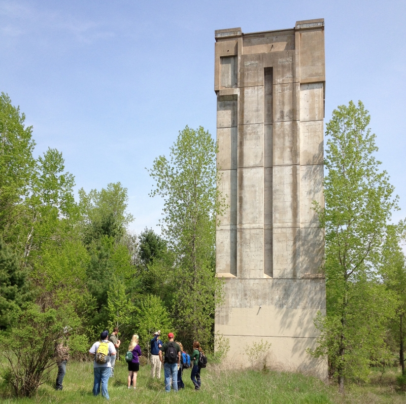 A picture of the Kickapoo Valley Reserve dam tower showing a group of people before a concrete tower rising nearly 100 feet above a grassy lawn
