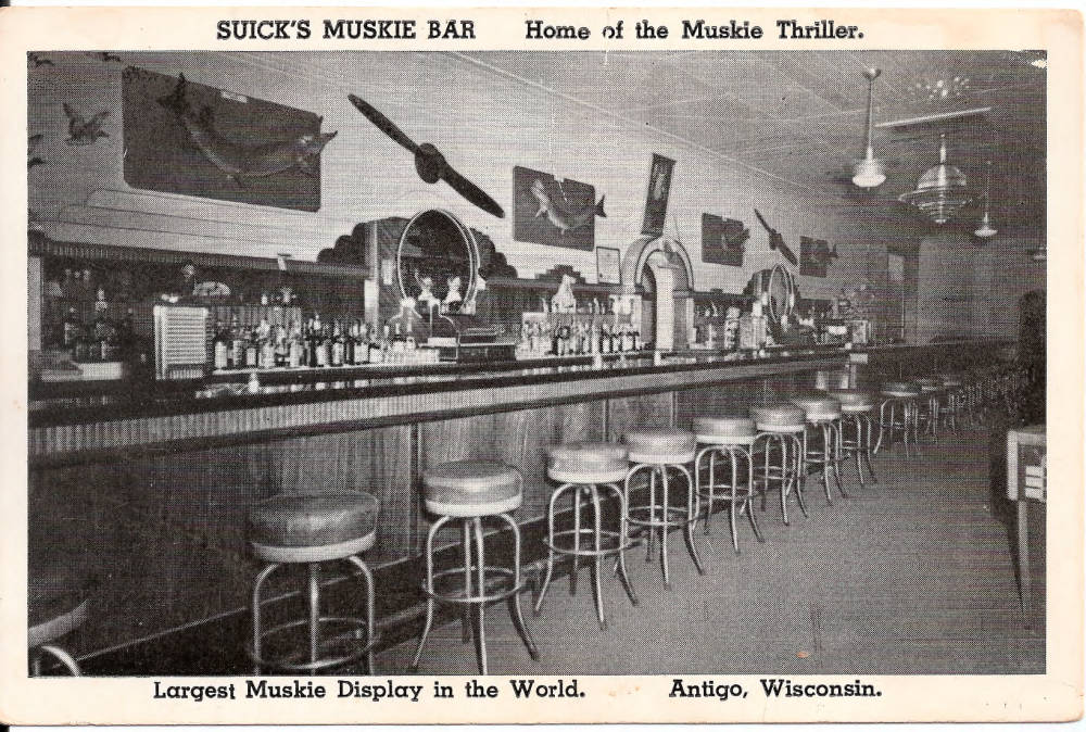 Postcard from Suick's Muskie Bar with photo of empty bar with large fish mounts on the wall. Text reads, Suick's Muskie Bar, Largest Muskie Display in the World.