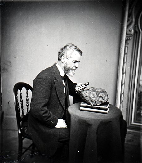 A posed photo portrait of Increase Lapham sitting at a desk with a magnifying glass examining a rock.