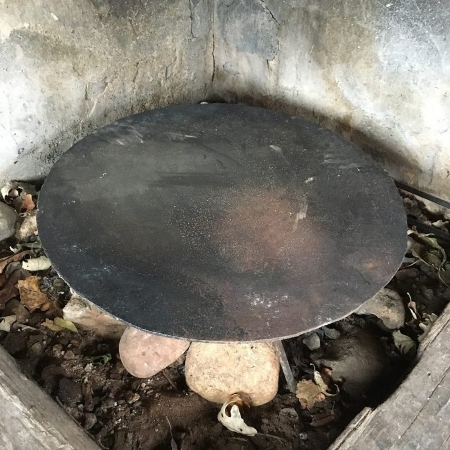 A round black griddle sitting atop stones in an open fireplace.
