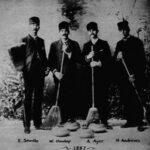 A black and white photograph of four men in suits posed with straw booms and curling stones.