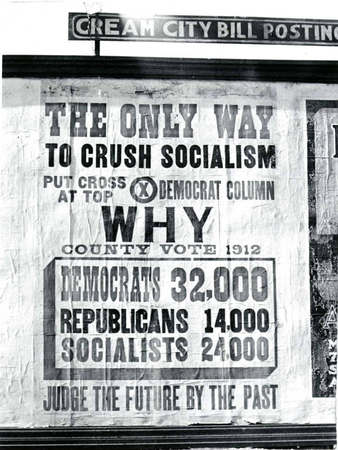 A black-and-white photograph of a wheat-paste poster advertisement against socialism on a building. The advertisement reads, "The only way to crush Socialism, put cross at top Democrat column. Why County Vote 1912. Democrats 32,000, Republicans 14,000, Socialists, 24,000. Judge the future by the past.