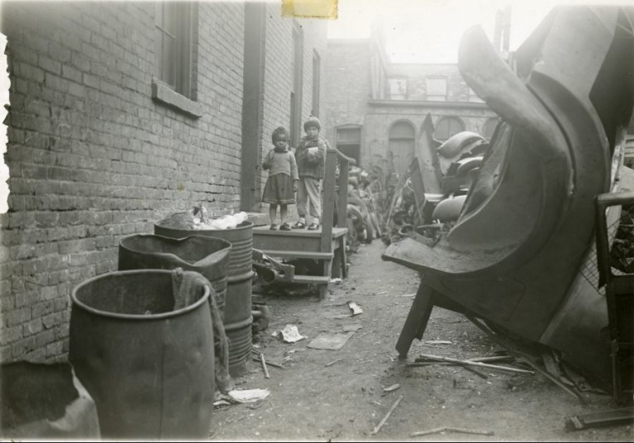 This photograph shows the rear of a building on N 8th St. Scrap metal from junked cars fills the yard. Two small children stand on a porch, staring at the camera.