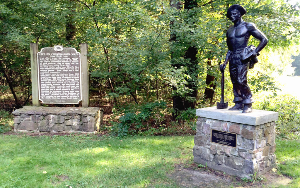 A historical plaque and statue of a shirtless man leaning against a shovel in a park setting.