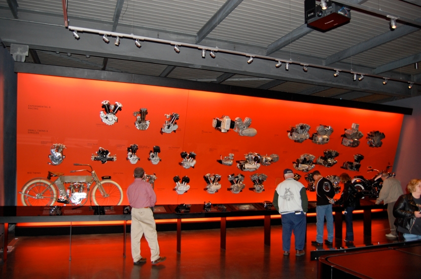 An image of museum visitor looking at motorcycle parts displayed on a wall, lit in red.
