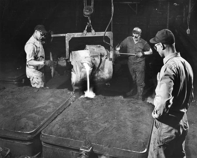 Photograph of three men pouring molten iron from a large cauldron into a sand mold.