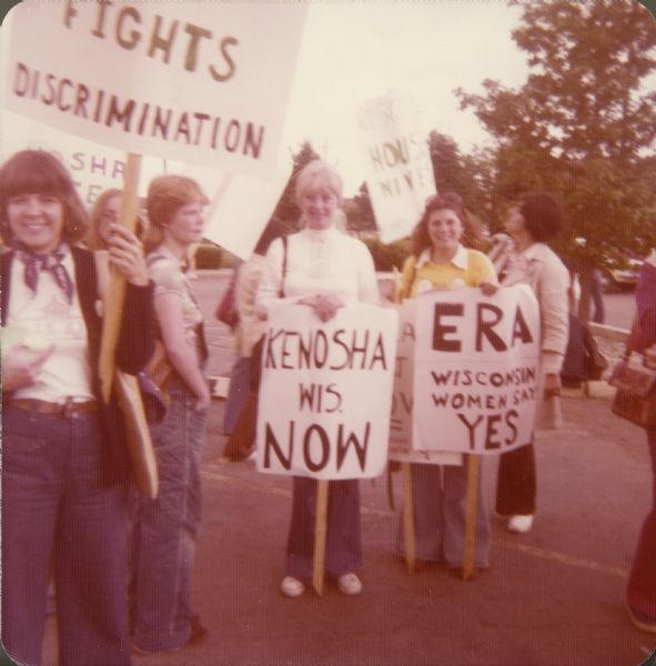 A group of women are standing outdoors holding signs. One sign reads: "Fights Discrimination." Other signs read: "Kenosha Wis. NOW" and "ERA Wisconsin Women Say YES,"