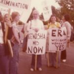 A group of women are standing outdoors holding signs. One sign reads: "Fights Discrimination." Other signs read: "Kenosha Wis. NOW" and "ERA Wisconsin Women Say YES,"