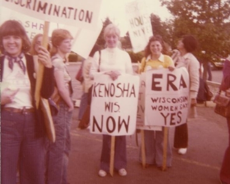 A group of women are standing outdoors holding signs. One sign reads: "Fights Discrimination." Other signs read: "Kenosha Wis. NOW" and "ERA Wisconsin Women Say YES,"