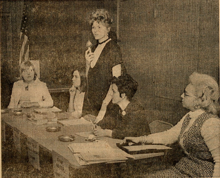 A newspaper photo clipping of five women at a table in front of a crowd. One woman stands with a microphone to address the crowd.