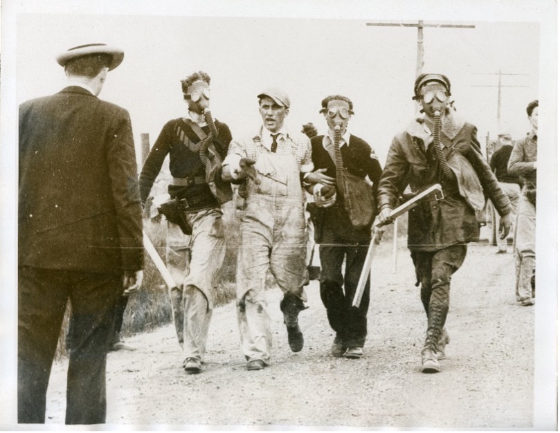 A photograph focused on four men walking down the street. Three wear gas masks and carry nightsticks. They hold another man by his arms, guiding him down the street.