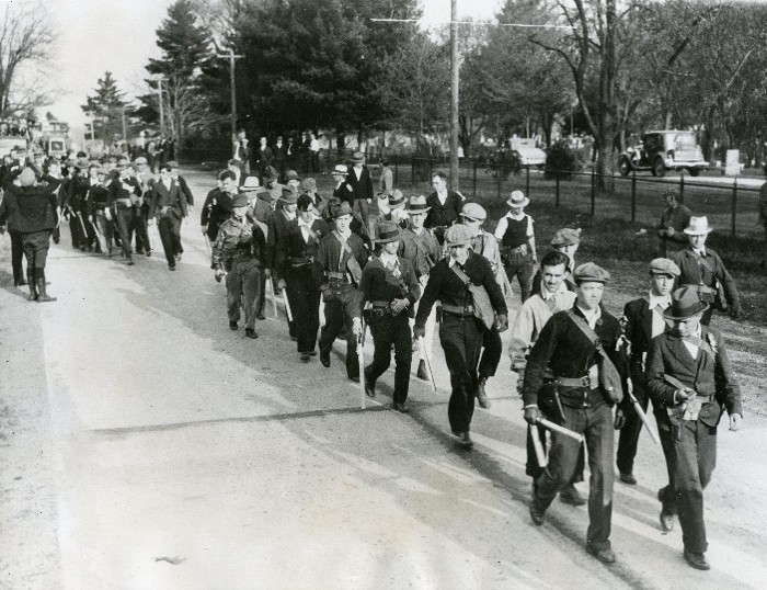 A group of men in various state of uniform march down a street in something of a makeshift parade.