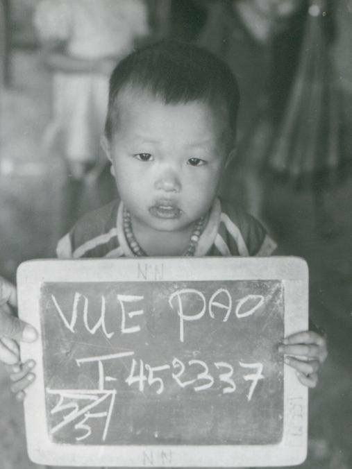 A black and white image of the author as a young boy in a Thai refugee camp holding a sign with identifying information.
