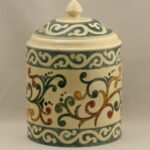 Hand-painted lidded jar. Shaped like a can with a slightly domed lid with finial. The base is cream color, and the painted ornament is of green, red, yellow, and blue vines.