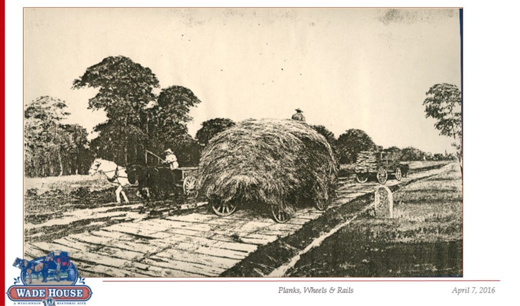 A historic image of horse-drawn wagons travelling along a plank road, hauling hay and logs.