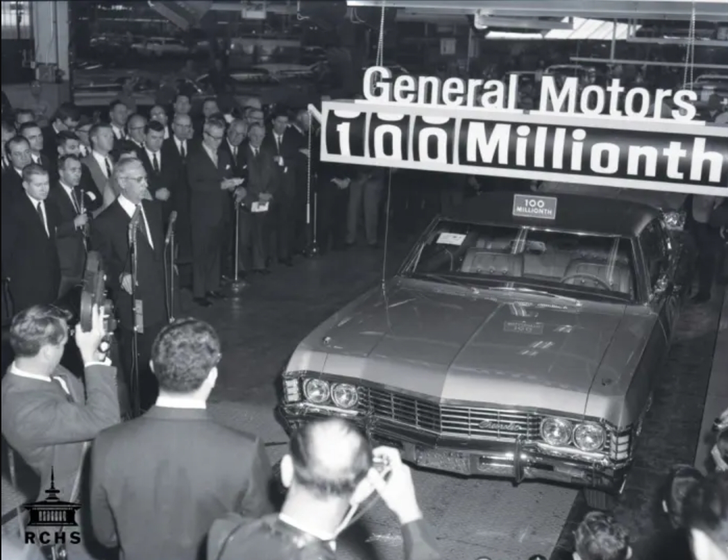 A crowd of people in a factory gather around a car at the end of an assembly line. A sign above reads, "General Motors 100 Millionth."