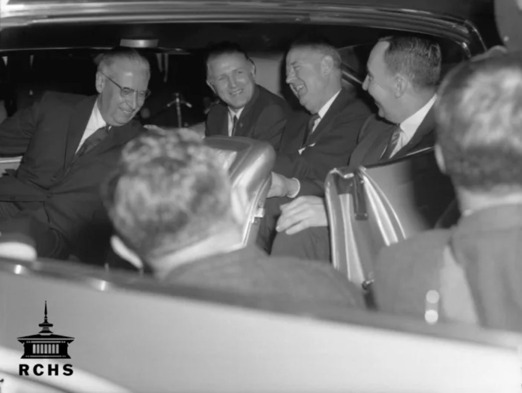 Four men in suits photographed laughing inside a car.