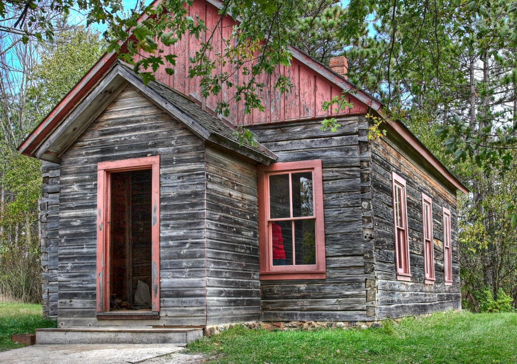 A one-room schoolhouse constructed of logs. Door and window frames are painted red.