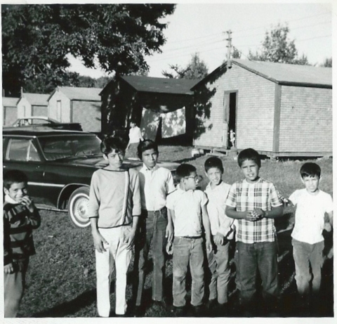 A group of children pose in a labor camp. Behind them is a car and a line of identical small buildings.