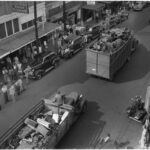 Trucks filled with scrap materials drive down a main street as people watch from the sidewalk.