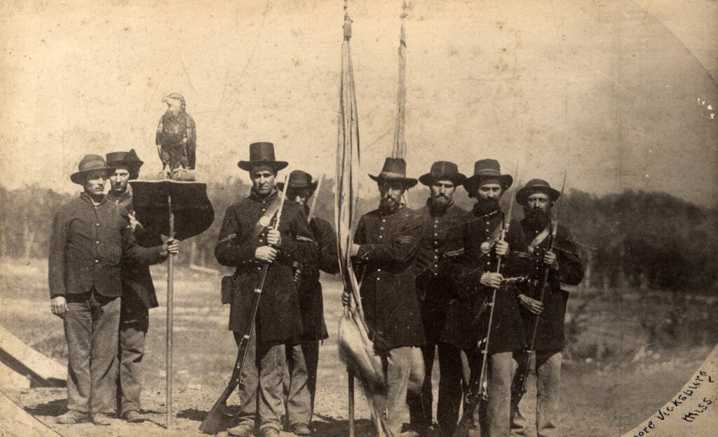 A photograph of eight Union soldiers, two hold flags, four hold rifles, and one holds a wooden pole with a shield-shaped wooden perch, atop which is an eagle.