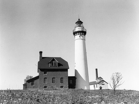 Black-and-white photograph of Outer Island Lighthouse and two-and-a-half story brick keepers house. The lighthouse towers over the light keeper house. It is round, constructed of brick, stuccoed, painted white, and has an observation deck that encircles the lantern.