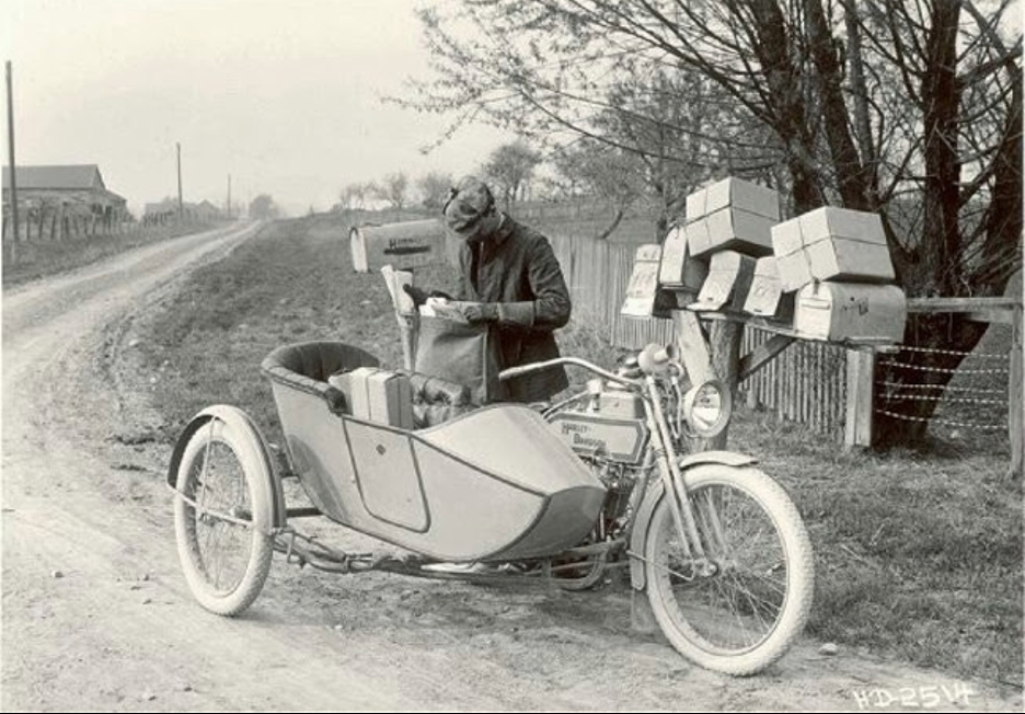Black-and-white photograph of a postman pulling parcels out of a sidecar attached to a motorcycle.