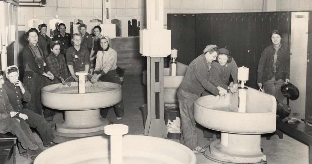 A group of women in welder’s gear pose around wash sinks inside a locker room.