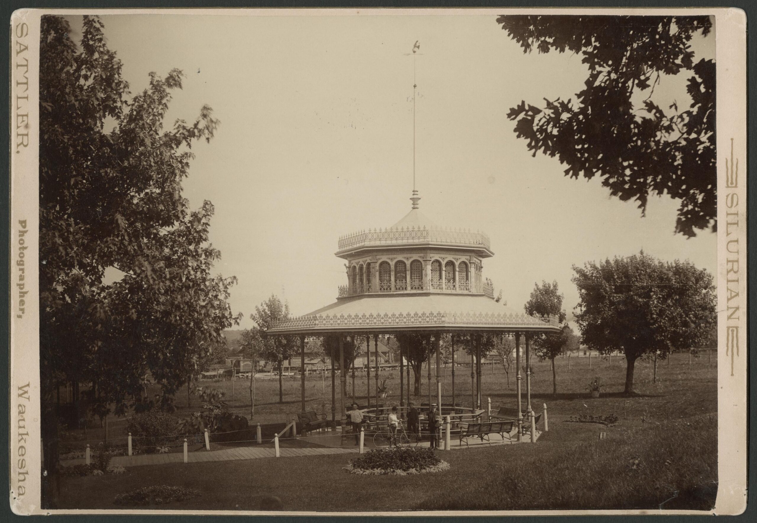 An ornate gazebo with stained glass windows and decorative roofing shelters the spring. People stand in the springhouse. One man has a bicycle, and another man holds drinking mugs.