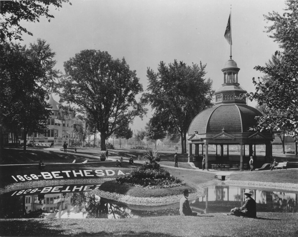 Black-and-white photograph of a spring water park, with a pool in the foreground, a pavilion in the midground, and a large hotel building in the background.
