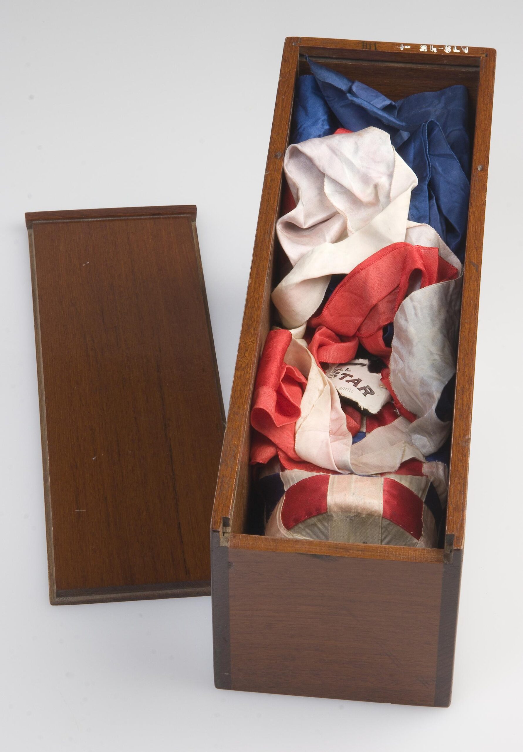 A small wooden box filled with red, white, and blue fabrics. The butt of a bottle, encased in woven red and white silk ribbons is visible at the bottom of the box.