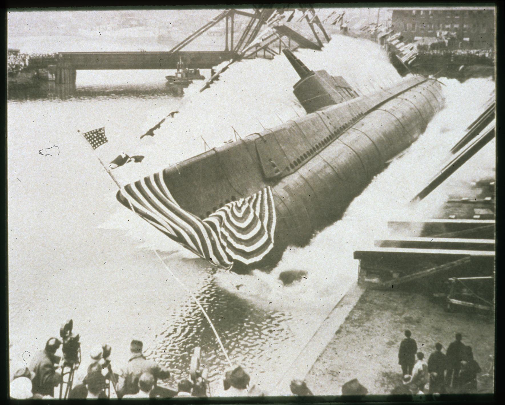 A warship submarine is launched into a harbor as crowds of onlookers watch.