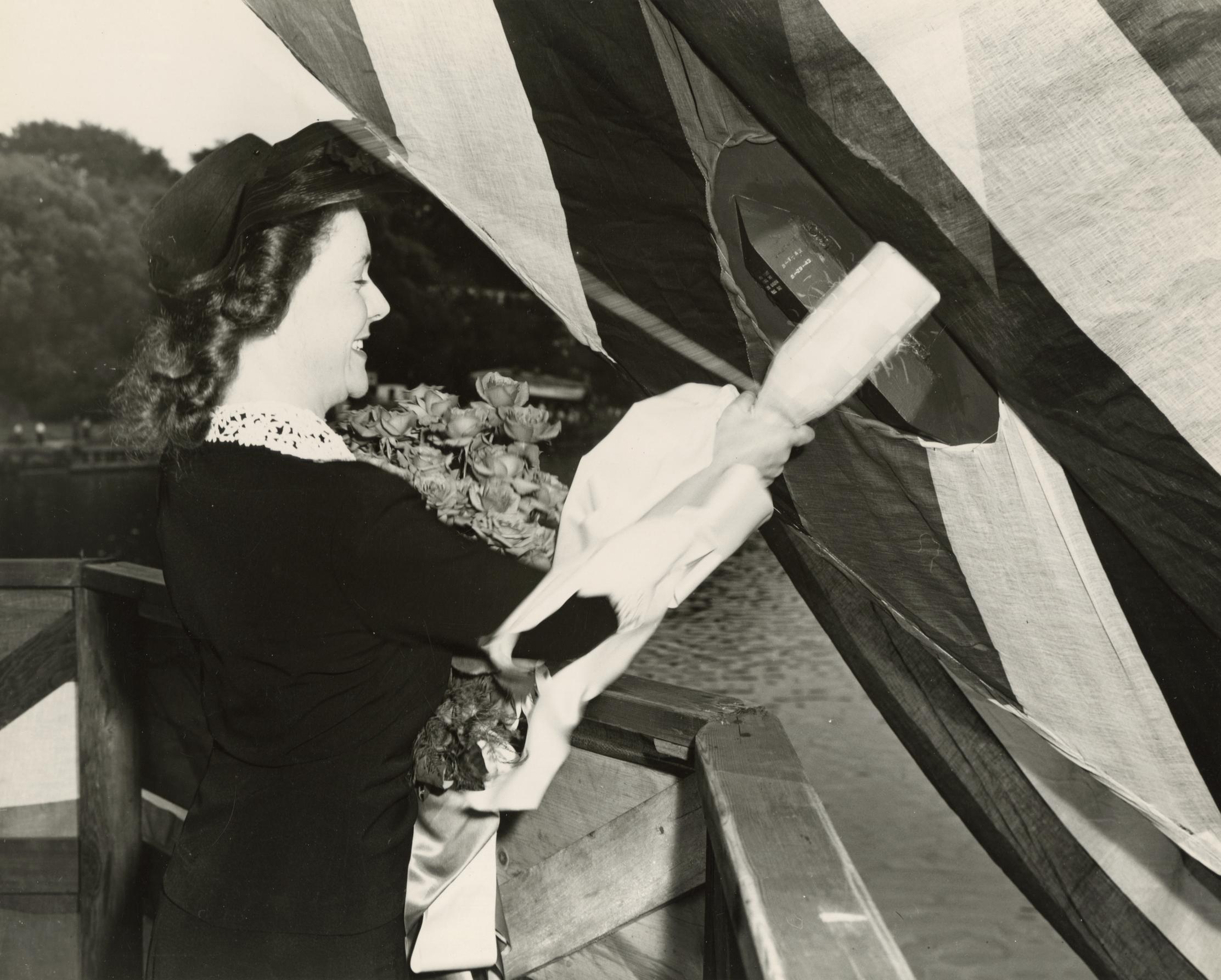 A black-and-white photograph of a young woman, cradling a bouquet of roses in one arm, and using the other to strike a decorated bottle against the bow of a warship.
