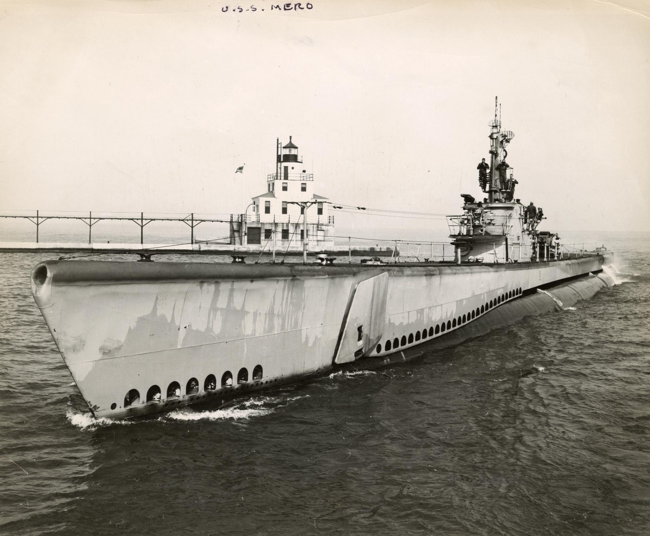 A large warship entering the harbor. About a dozen crewmen and officers are seen on the bridge and platform around the conning tower.