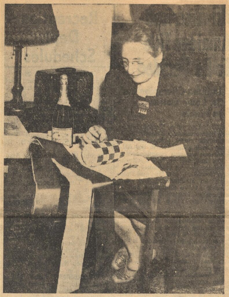 A photograph of a woman, seated in a home setting, sewing ribbons around a bottle.