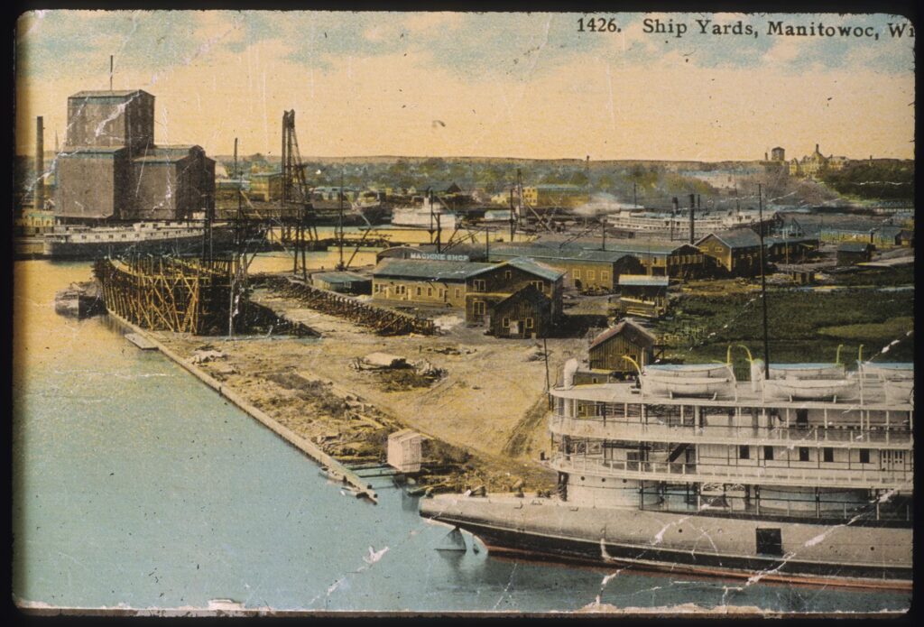 A colorized postcard of the ship yards at Manitowoc. At the far left there is a grain elevator and large cargo ship. Also visible are many warehouses and large shop buildings, and a ship in scaffolding wither being built or repaired.