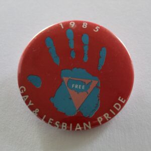 A red button pin which features a blue handprint, in the palm of which is an inverted pink triangle and an image of the United States. Text around the border reads "Gay & Lesbian Pride, 1985"