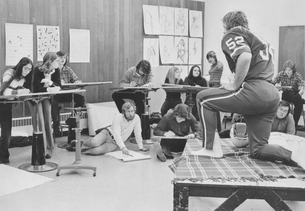 A photograph of a life drawing class where art students gather around a live male model dressed in football uniform.