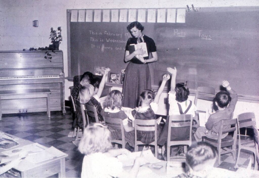 A young woman stands in front of a group of children in a classroom gesturing at a book as children attentively raise their hands.