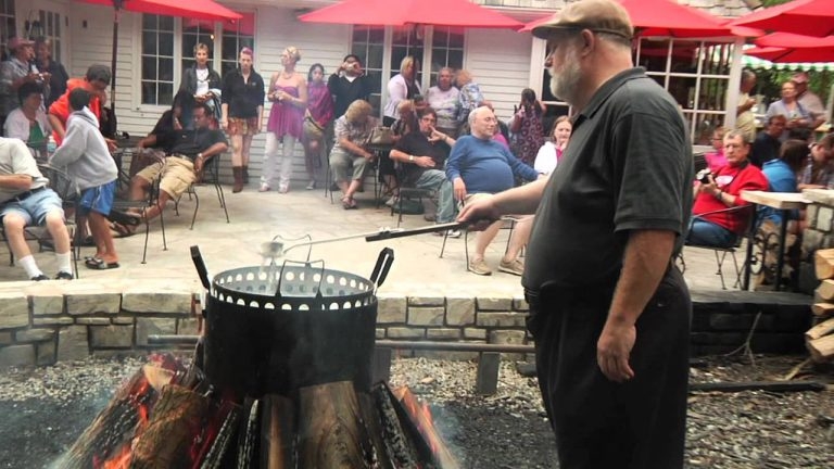 A man holds a two-sided spork and tends a boil pot on an open fire. A crowd of people watch in the background.