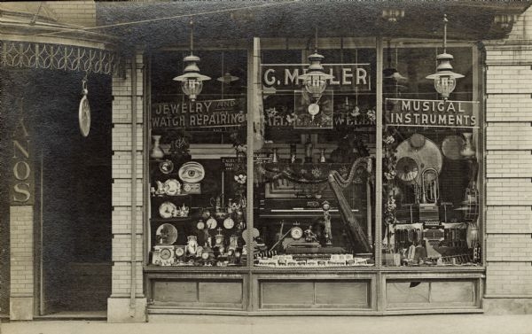 A black-and-white photograph of a storefront display featuring a variety of musical instruments and clocks.