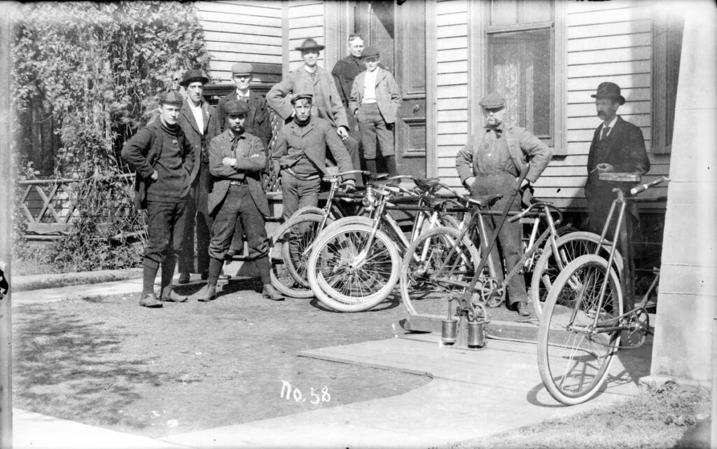 Group of men, and a young boy, standing in the small front yard of a house at a bicycling outing. A woman stands in an open doorway at the top of the steps. A man on the far right wears a suit and appears to be holding a cigar. Bicycles are leaning against a railing in front of the house. The men wear sweaters, jackets, hats, stockings, button-up shoes and knickerbockers. On top of a wood sidewalk in the yard is what appears to be a hand-pump with a long handle for putting air into bicycle tires.