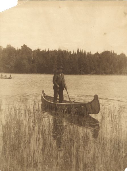 Sepia-tone photograph across water of a Menominee man standing in a canoe in a marsh. He is holding a paddle in his hand. Behind him on the left is another canoe with three people. The far shoreline is lined with trees.