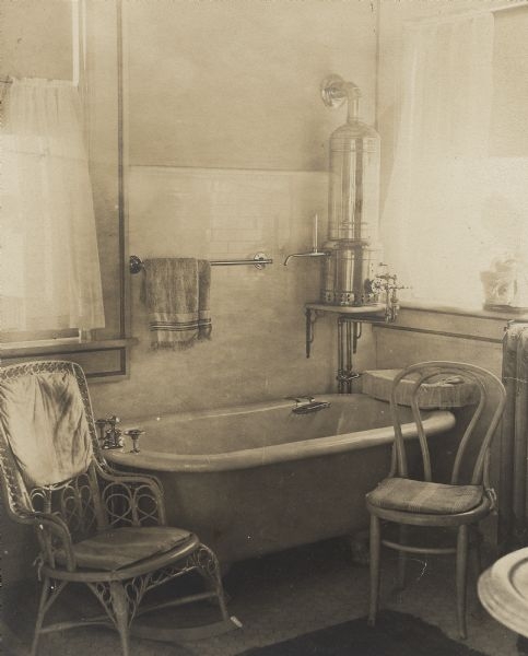A wicker rocker and bentwood side chair flank the bathtub in a bathroom of the Frank Brown Fargo house. There is a hot water heater above the tub in the corner of the room and a steam radiator is on the right. The floor and walls are tiled. Two windows fill the room with light.