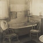 A wicker rocker and bentwood side chair flank the bathtub in a bathroom of the Frank Brown Fargo house. There is a hot water heater above the tub in the corner of the room and a steam radiator is on the right. The floor and walls are tiled. Two windows fill the room with light.