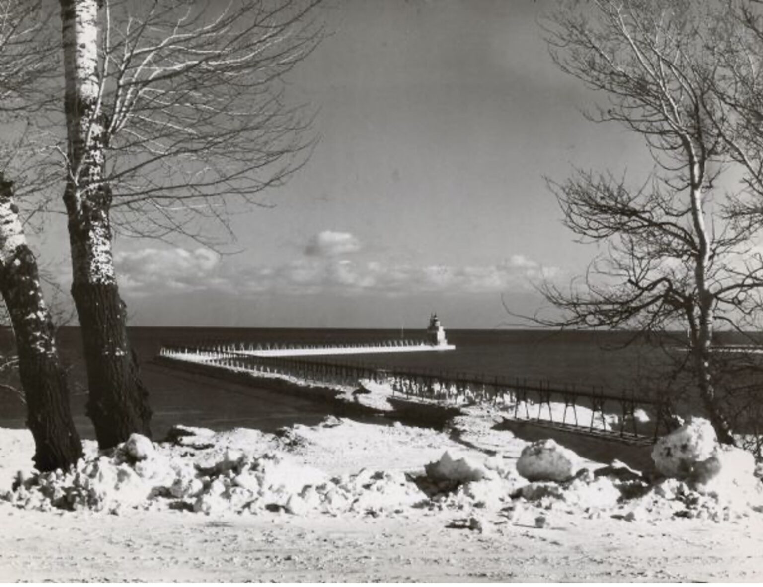 A view of the breakwater and lighthouse from the shore. Snow and trees are along the Lake Michigan shoreline in the foreground.