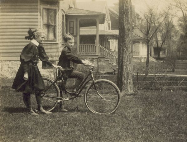 A boy mounts a bicycle in the yard, his sister steadies the bike by holding onto the seat.