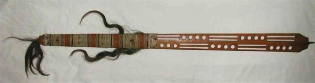A wooden pipe stem, flat, half-carved with circles and rectangles, and decorated with quillwork in white, orange, red, and brown, and horsehair.